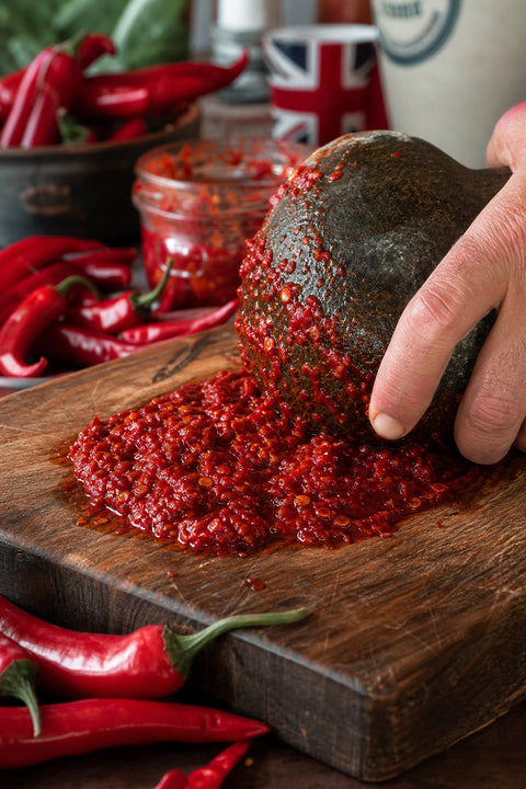 A close‑up of fresh red chili peppers being ground into a rustic paste on a wooden cutting board, natural lighting, real